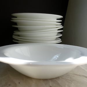 May include: A stack of white ceramic bowls, with a single bowl in the foreground. The bowls are all the same size and shape, and they are arranged in a neat pile.