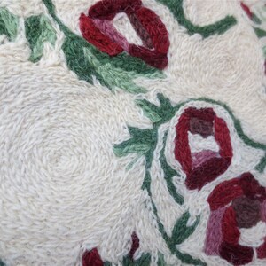 K&ouml;nnte beinhalten: Nahaufnahme eines wei&szlig;en Stoffes mit roten und gr&uuml;nen gestickten Blumen. Die Blumen sind in einem kreisf&ouml;rmigen Muster angeordnet und haben ein detailliertes Design.