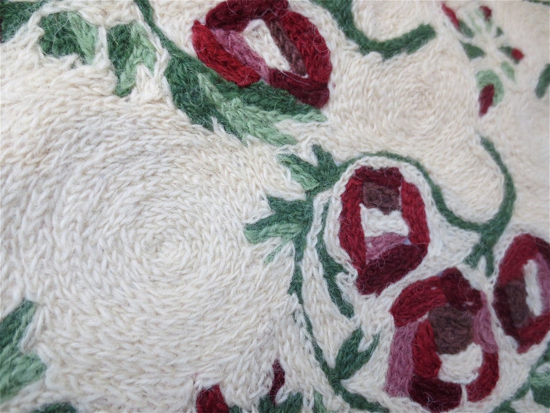 K&ouml;nnte beinhalten: Nahaufnahme eines wei&szlig;en Stoffes mit roten und gr&uuml;nen gestickten Blumen. Die Blumen sind in einem kreisf&ouml;rmigen Muster angeordnet und haben ein detailliertes Design.