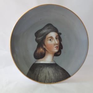 May include: A round decorative porcelain plate featuring a hand-painted portrait of a person with shoulder-length brown hair wearing a dark cap and a black garment. The background is a solid grey, and the plate has a thin gold-colored rim.