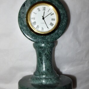 May include: A green marble quartz clock with a gold-colored bezel and white face. The clock has Roman numerals and black hands. The clock is mounted on a green marble base and pedestal.
