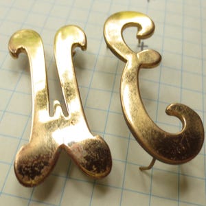 May include: Two vintage gold-toned metal letter-shaped ornaments. One is a stylized "W" with a central bar, and the other is a cursive "E". Both have a decorative, scrolled design. The ornaments are on a grid background.