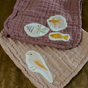 Op de afbeelding: Twee vierkante mousseline doeken met een gekreukte textuur. De bovenste doek is oudroze met patches van een heremietkreeft, een kempvis en een goudvis. De onderste doek is perzikkleurig en heeft een patch van een vogel met gele accenten.