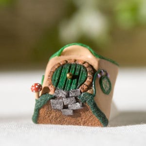 Peut inclure: Figurine miniature représentant une maison de hobbit avec une porte verte, des marches en pierre et un champignon rouge. La maison a un extérieur beige avec des accents verts et un toit vert. Une petite pièce décorative détaillée.