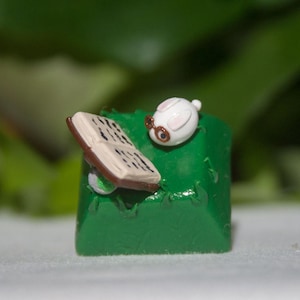 May include: A green keycap featuring a white rabbit figurine reading a book. The rabbit has brown eyes and a brown nose.