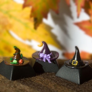 May include: Three miniature witch hats on black bases. The hats are made of felt and have different colours and designs. One hat is purple with a crescent moon, one is brown with green accents, and one is black with a gold buckle.