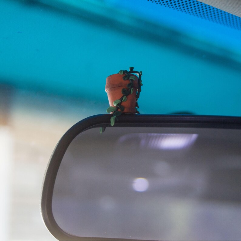 Miniature potted ivy plant monitor topper made out of polymer clay. The plant is dark green with some lighter green leaves. The pot is terra cotta. This figure is on top of a car rearview mirror.