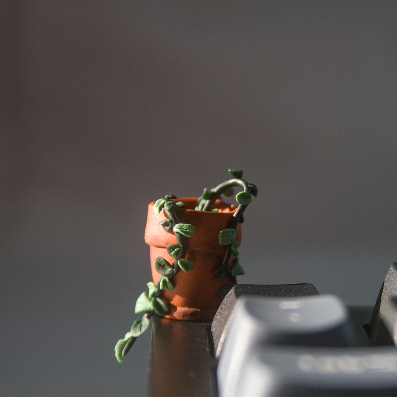 Miniature potted ivy plant monitor topper made out of polymer clay. The plant is dark green with some lighter green leaves. The pot is terra cotta. This figure is on top of a keyboard at the corner edge.
