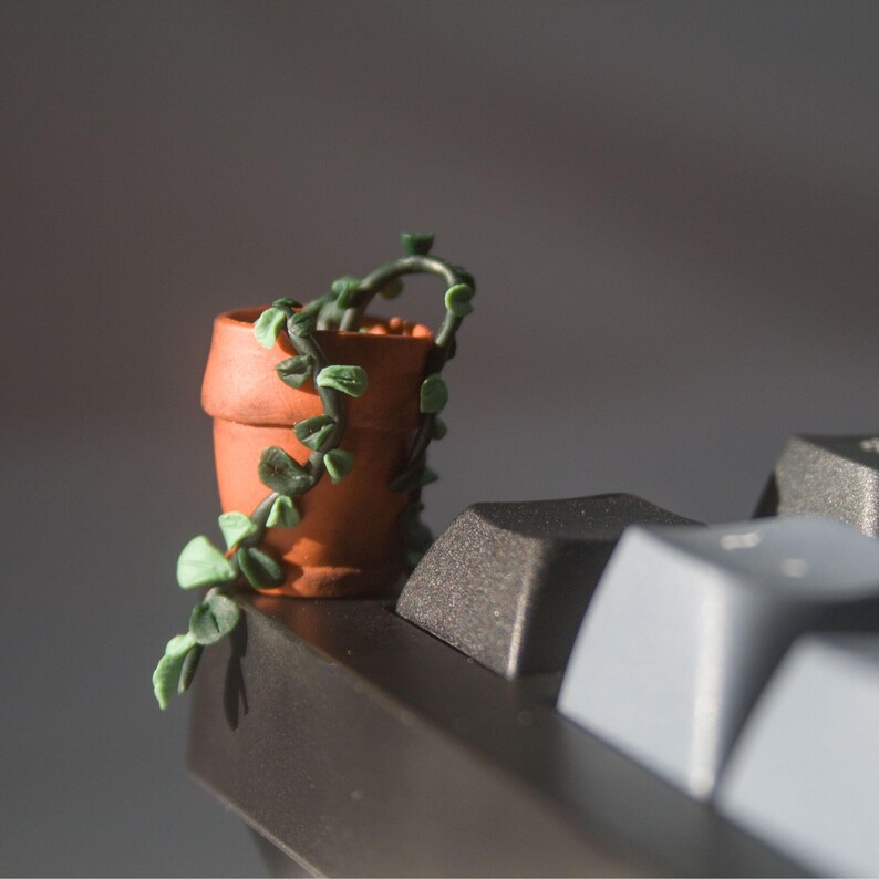 Miniature potted ivy plant monitor topper made out of polymer clay. The plant is dark green with some lighter green leaves. The pot is terra cotta. This figure is on top of a keyboard at the corner edge.
