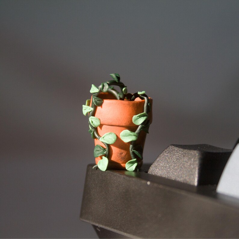 Miniature potted ivy plant monitor topper made out of polymer clay. The plant is dark green with some lighter green leaves. The pot is terra cotta. This figure is on top of a keyboard at the corner edge.