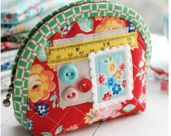May include: A red quilted pouch with a green and white checkered trim. The pouch has a zipper closure and is decorated with buttons, a ruler, and a fabric flower patch. The pouch is a sewing notions pouch.