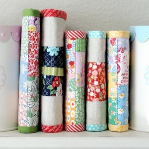 May include: Five fabric-covered journals with colourful patchwork designs. The journals are lined up in a row on a white surface. The journals are all different colours and patterns, including red, green, blue, and yellow.