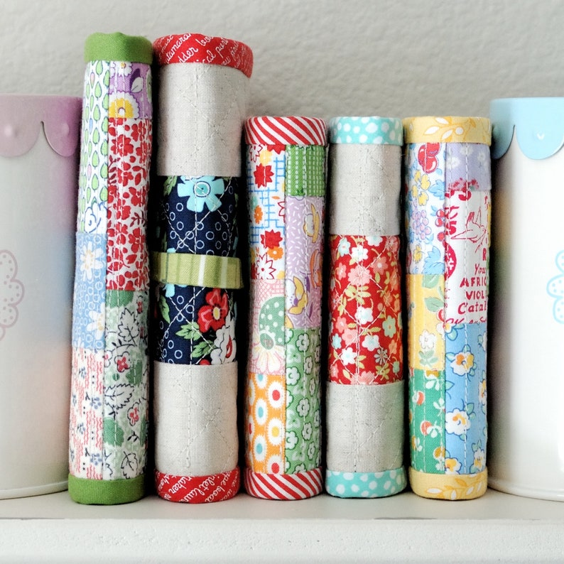 May include: Five fabric-covered journals with colourful patchwork designs. The journals are lined up in a row on a white surface. The journals are all different colours and patterns, including red, green, blue, and yellow.