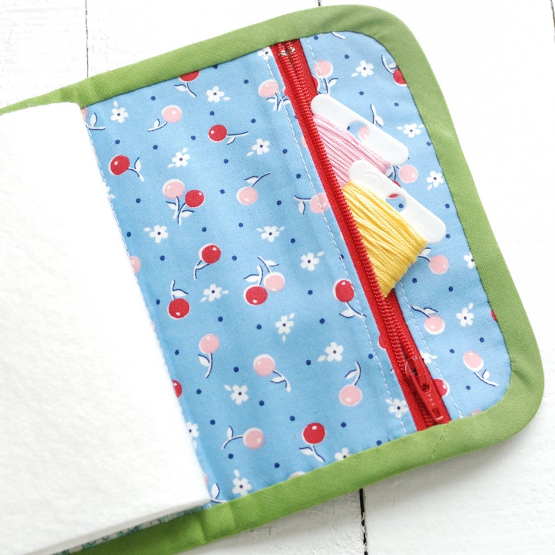 May include: A green fabric zippered pouch with a blue and white cherry print lining. The pouch is open and has three spools of embroidery floss inside.