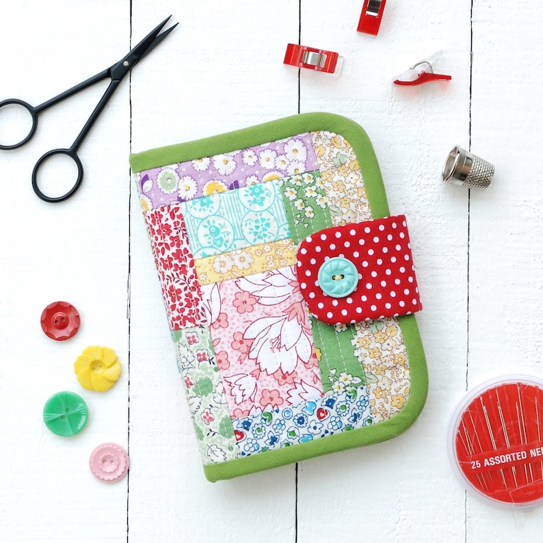 May include: A patchwork fabric sewing kit with a red and white polka dot flap, a turquoise button, and a green border. The kit is on a white wooden surface with sewing supplies.