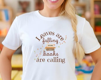 May include: White t-shirt with the text "Leaves are falling, books are calling" in brown lettering. The design includes illustrations of books and autumn leaves in shades of orange, green, and yellow. The shirt is a casual, short-sleeved style.