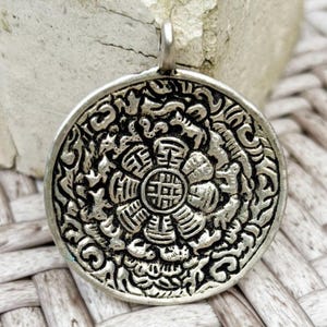 May include: A silver-colored pendant with an intricate floral and geometric design. The pendant has a loop at the top for a chain. The design features a central flower-like shape surrounded by detailed patterns and symbols. The pendant is likely made of metal.