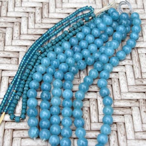 May include: A collection of blue bead strands, showcasing diverse shades and sizes. The beads vary from small, cylindrical forms to larger, spherical shapes. The strands are strung together, with some combining different bead types. The backdrop is a light-toned woven pattern.