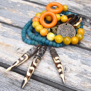 May include: A collection of handcrafted jewelry and decorative items. The image features a variety of beads in shades of blue, yellow, and orange, along with carved wooden pendants and a circular pendant with an engraved design. The items are arranged on a weathered wooden surface.
