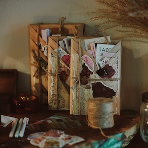 May include: Three brown paper packages tied with twine, decorated with vintage ephemera and wax seals. A white vase with dried flowers, a lit candle in a glass jar, and a wooden box are also visible.