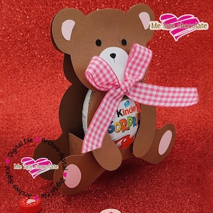Puede incluir: Un oso de peluche marrón con orejas rosas y una cinta de cuadros rosa y blanca alrededor del cuello. El oso sostiene un huevo de chocolate Kinder Surprise en sus patas. El texto "Me late chocolate" está escrito en rosa sobre un corazón blanco en la esquina superior derecha de la imagen.