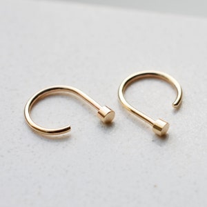 May include: A pair of gold-toned hoop earrings, each featuring a smooth, curved wire design that terminates in a small, flat, cylindrical stud at one end. The earrings have a polished, reflective finish and a simple, open-ended circular shape.