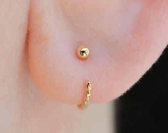 以下が含まれることがあります： 小さな滑らかで光沢のある球体を特徴とするゴールドのスタッドピアスと、ねじれた質感のデザインのゴールドのフープピアス。どちらも明るく反射するイエローメタルで作られており、耳の2つのピアスホールに装着されています。
