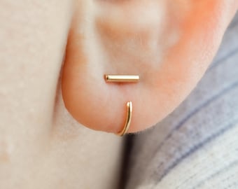 Op de afbeelding: Een gouden staafje als oorsteker horizontaal op de oorlel geplaatst, vergezeld door een dunne gouden oorring die rond de onderrand van de oorlel buigt, wat een gelaagde uitstraling creëert met twee afzonderlijke gouden sieraden.