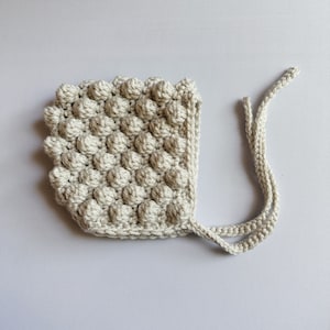 May include: A light beige, handmade baby bonnet with a textured, bobble stitch design. The bonnet has a rounded shape with a square edge and ties. The neutral color and soft texture suggest a cozy, handmade item, perfect for a newborn.