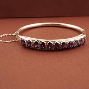 May include: A silver bangle bracelet with oval-cut purple gemstones. The bracelet has a hinged design and a safety chain. The gemstones are set in a row along the front. The background is a solid brown colour.
