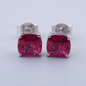 May include: A pair of square-cut, ruby-red gemstone stud earrings. The earrings have a silver-coloured setting and post. The gemstones are a vibrant, saturated pink-red colour. The earrings are displayed on a light blue background.