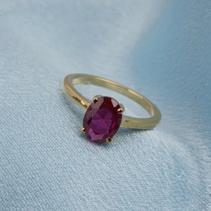 Könnte beinhalten: Ein goldener Ring mit einem ovalen, tiefroten Edelstein. Das Band des Rings ist aus poliertem Gold, und der Edelstein wird von vier goldenen Krappen gehalten. Der Ring ist vor einem hellblauen Stoffhintergrund platziert.