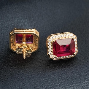 May include: A pair of gold earrings featuring a square red gemstone surrounded by smaller white gemstones.