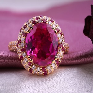 May include: A gold ring featuring a large, oval-shaped, deep pink gemstone surrounded by smaller, clear and pink gemstones.