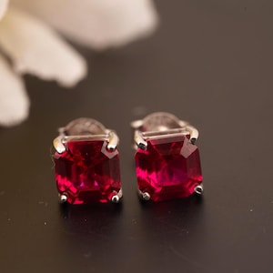 Pode incluir: Um par de brincos de botão com pedras preciosas vermelho rubi de corte quadrado e engastes prateados. Os brincos estão colocados sobre um fundo escuro, com uma flor branca desfocada no canto superior esquerdo.