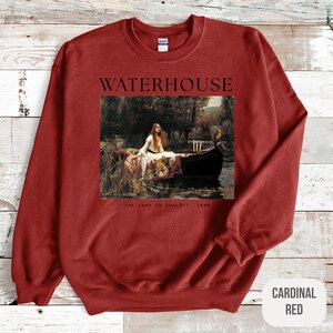 May include: Red sweatshirt with a painting of a woman in a boat on the front. The painting is titled "The Lady of Shalott, 1888" and the artist's name, "WATERHOUSE", is printed above the painting.