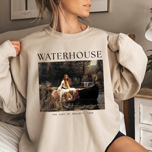 May include: A beige sweatshirt with a graphic print of a painting by John William Waterhouse titled "The Lady of Shalott" from 1888. The text "WATERHOUSE" is printed above the painting.