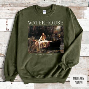 May include: A military green sweatshirt with a white print of a painting by John William Waterhouse titled "The Lady of Shalott, 1888". The painting depicts a woman in a boat on a river.