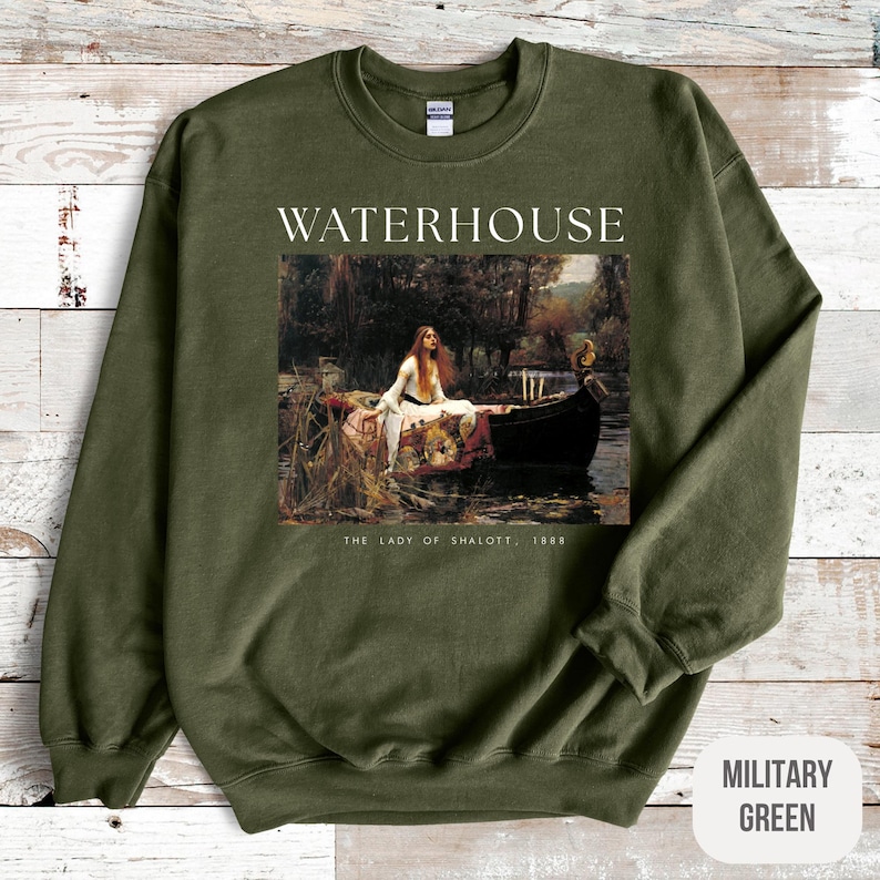 May include: A military green sweatshirt with a white print of a painting by John William Waterhouse titled "The Lady of Shalott, 1888". The painting depicts a woman in a boat on a river.