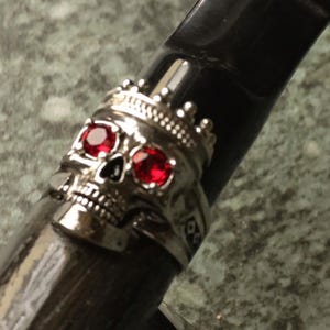Könnte beinhalten: Ein silberner Totenkopfring mit Krone und roten Edelsteinaugen ist auf einem dunklen, strukturierten Objekt abgebildet. Das Design des Rings weist komplizierte Details auf, und die Edelsteine ​​sorgen für einen Farbtupfer. Das Objekt scheint eine Pfeife oder ein ähnliches Objekt zu sein.