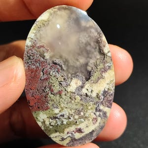 Könnte beinhalten: Ein polierter ovaler Cabochon aus Achat mit einem wirbelnden Muster aus Weiß, Grau, Grün und Rot.