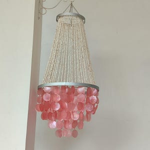 Könnte beinhalten: Ein rosa-weißer Muschel-Kronleuchter mit silbernem Rahmen. Der Kronleuchter besteht aus vielen kleinen, runden Muscheln, die aneinandergereiht sind.