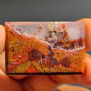 Może przedstawiać: Przejrzysty, prostokątny kaboszon agatowy z warstwowym wzorem w kolorze brązowym, pomarańczowym i czerwonym. Agat ma białe, zamglone centrum.