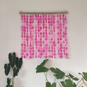 Puede incluir: Cortina de conchas rosadas hecha de conchas redondas que se superponen y cuelgan de una barra de madera. La cortina está colgada en una pared blanca.