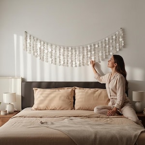 Peut inclure: Une décoration murale composée de multiples brins de disques de coquillages blancs translucides et circulaires suspendus à un cordon horizontal. Les disques sont disposés en un motif semi-circulaire en cascade, créant un aspect texturé et superposé.