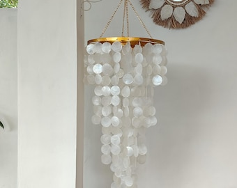 May include: A decorative chandelier with a gold-coloured metal frame and hanging strands of white, round shell pieces. The chandelier is suspended from a metal hook. A round mirror with a woven, sunburst-style frame hangs on the wall.