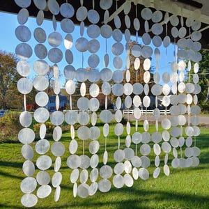 Può includere: Una tenda di conchiglie bianche composta da molte conchiglie rotonde e sovrapposte appese a una trave di legno bianco. La tenda è appesa davanti a un prato verde e un cielo blu.