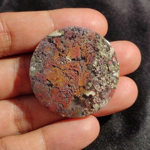 May include: A round, polished stone with a unique pattern of red, orange, purple, and green hues. The stone's surface has a textured appearance, with intricate details and variations in color. The stone is held in a hand.