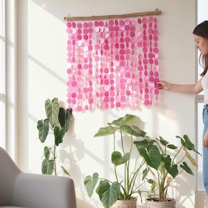 Peut inclure: Une décoration murale composée de multiples brins verticaux de disques roses circulaires suspendus à une branche de bois horizontale. Les disques varient dans les tons de rose et sont disposés en grille, créant un effet de rideau décoratif.