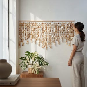 May include: A wall hanging composed of numerous circular, translucent, light brown shell discs suspended from a horizontal wooden rod. The discs are arranged in multiple vertical strands of varying lengths, creating a layered, textured appearance.